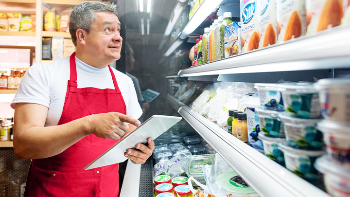 一名身穿红围裙的超市工作人员拿着平板电脑看着货架上的食品